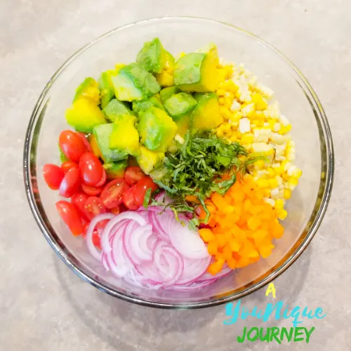 All the ingredients: corn, avocado, red onion, grape tomatoes, bell pepper and basil in a large glass bowl.