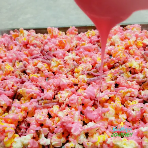 Pouring the remaining melted bright pink candy melts wafers over the coated popcorn and broken pretzels.